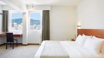 A hotel room with a bed, a desk, and a chair. There is a window with curtains and a view of buildings and hills.