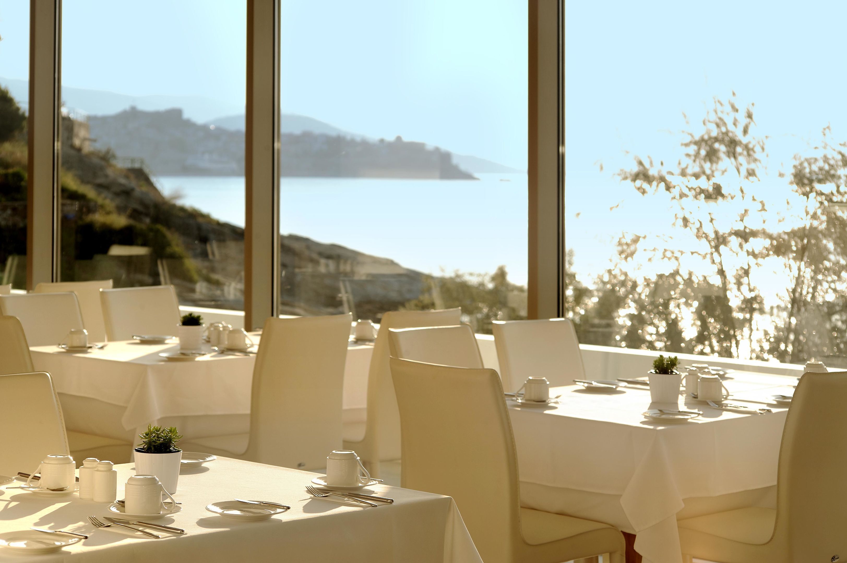 A restaurant with tables set for dining, large windows offering a view of the sea and hills.