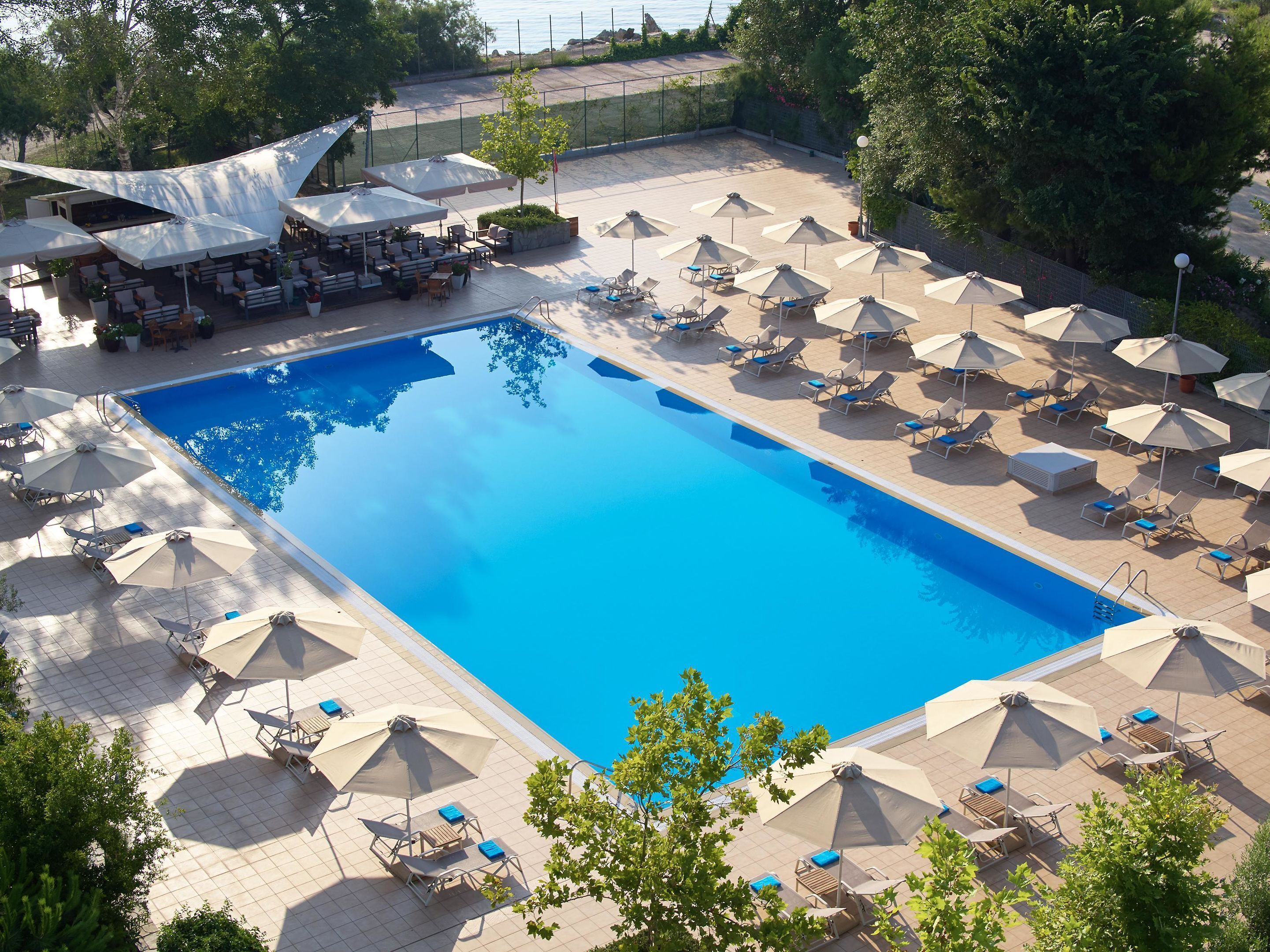 A large rectangular swimming pool surrounded by sun loungers and umbrellas, with a seating area and a covered section in the background.