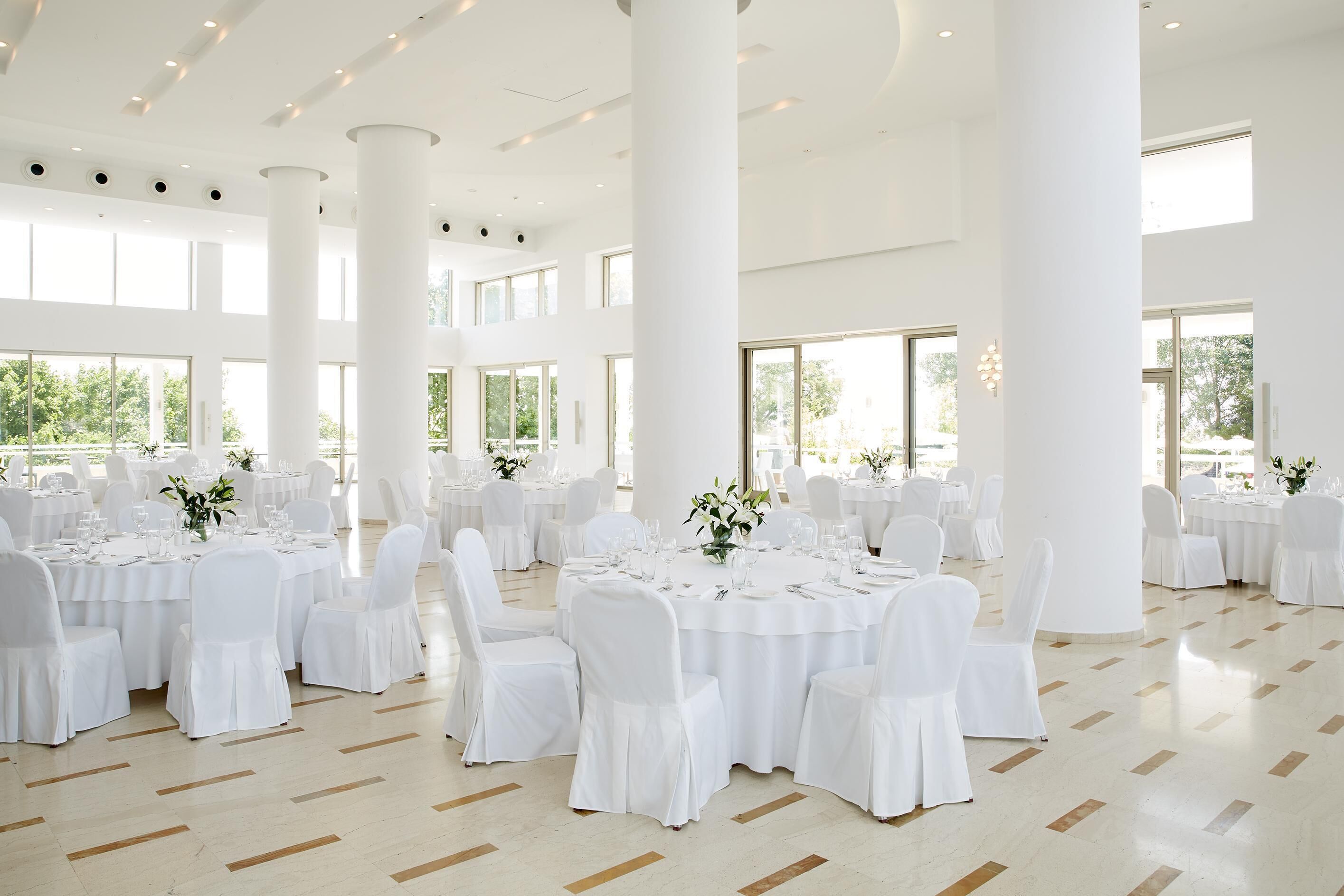 Un salón amplio con mesas y sillas blancas, ventanas grandes y un techo alto.