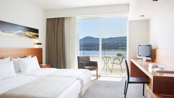 A hotel room with a large bed, a desk with a computer, a chair, and a view of a lake and mountains.