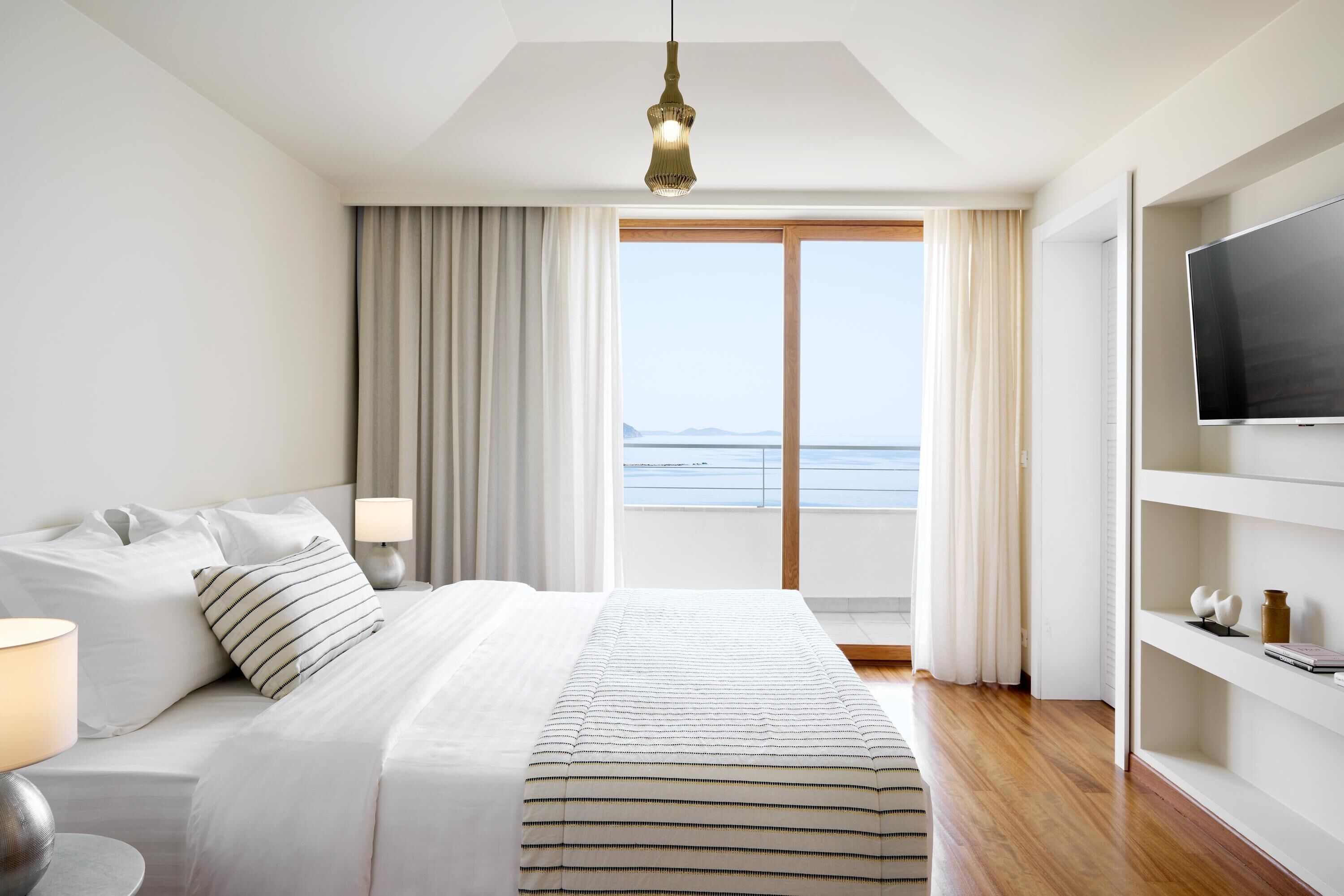 A hotel room with a bed, a TV, a bookshelf, and a view of the sea.