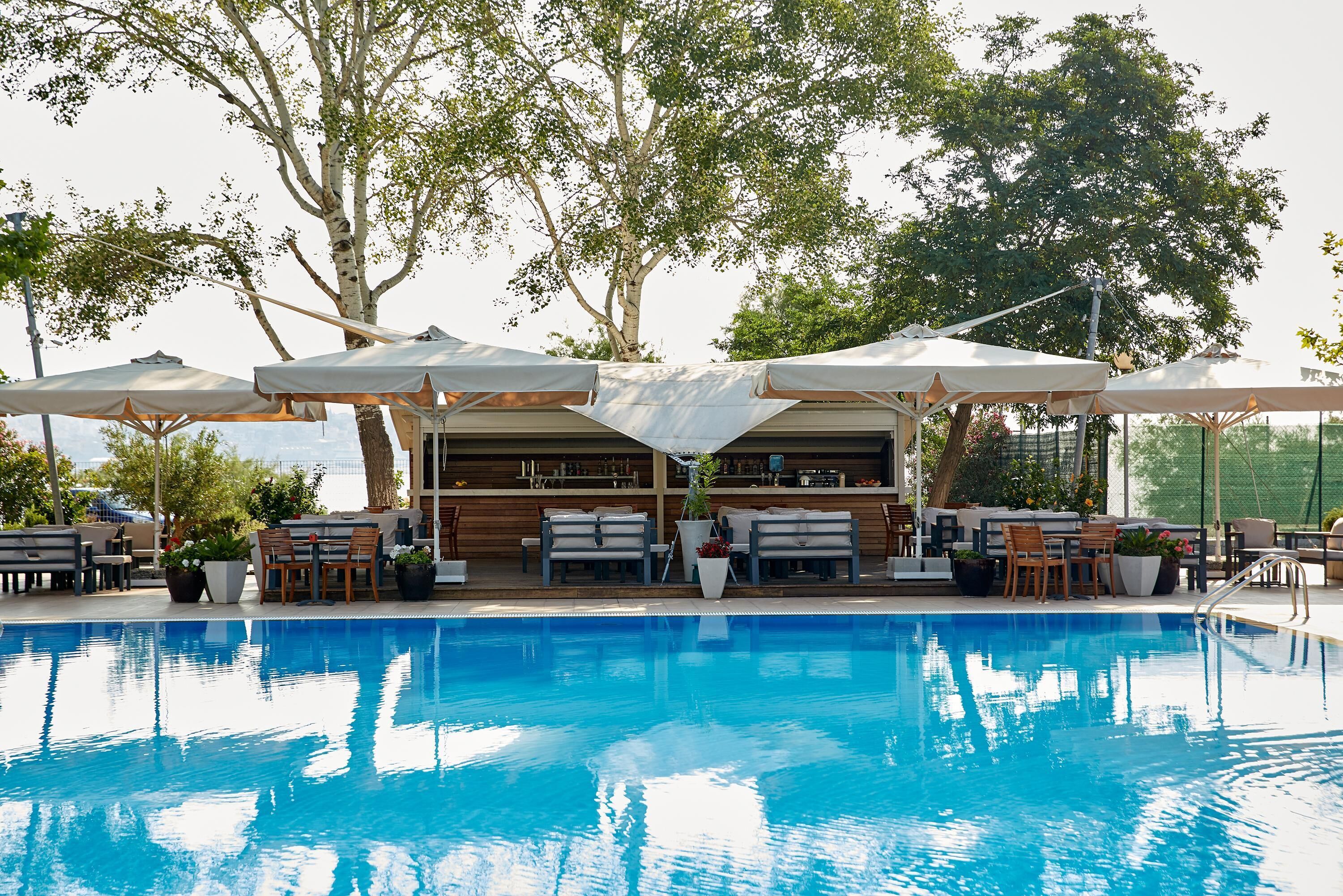 Zona de piscina amb gandules i ombrel·les, envoltada d'arbres i una zona de bar.