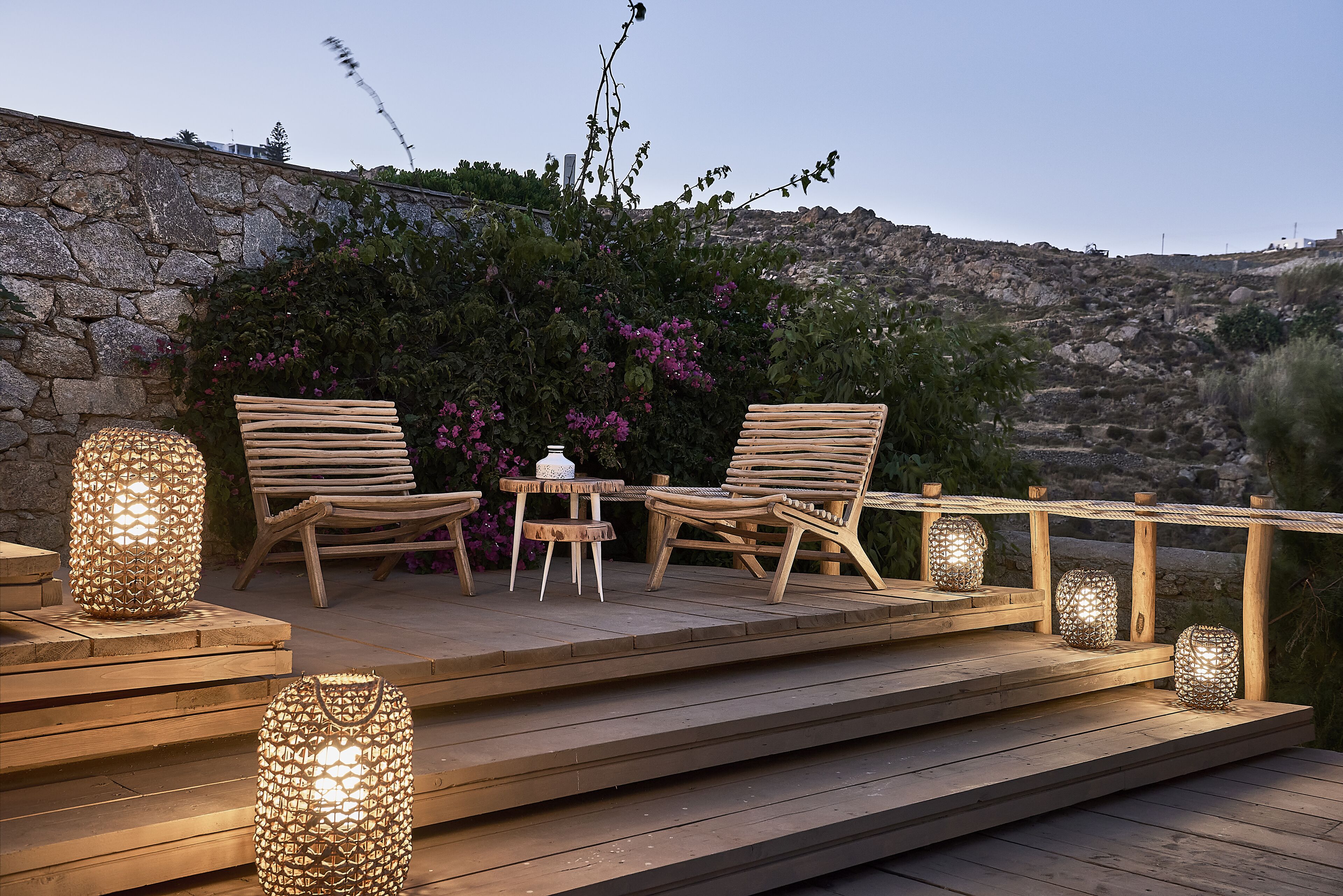 A wooden deck with two wicker chairs and a small table, surrounded by decorative lanterns and a stone wall.