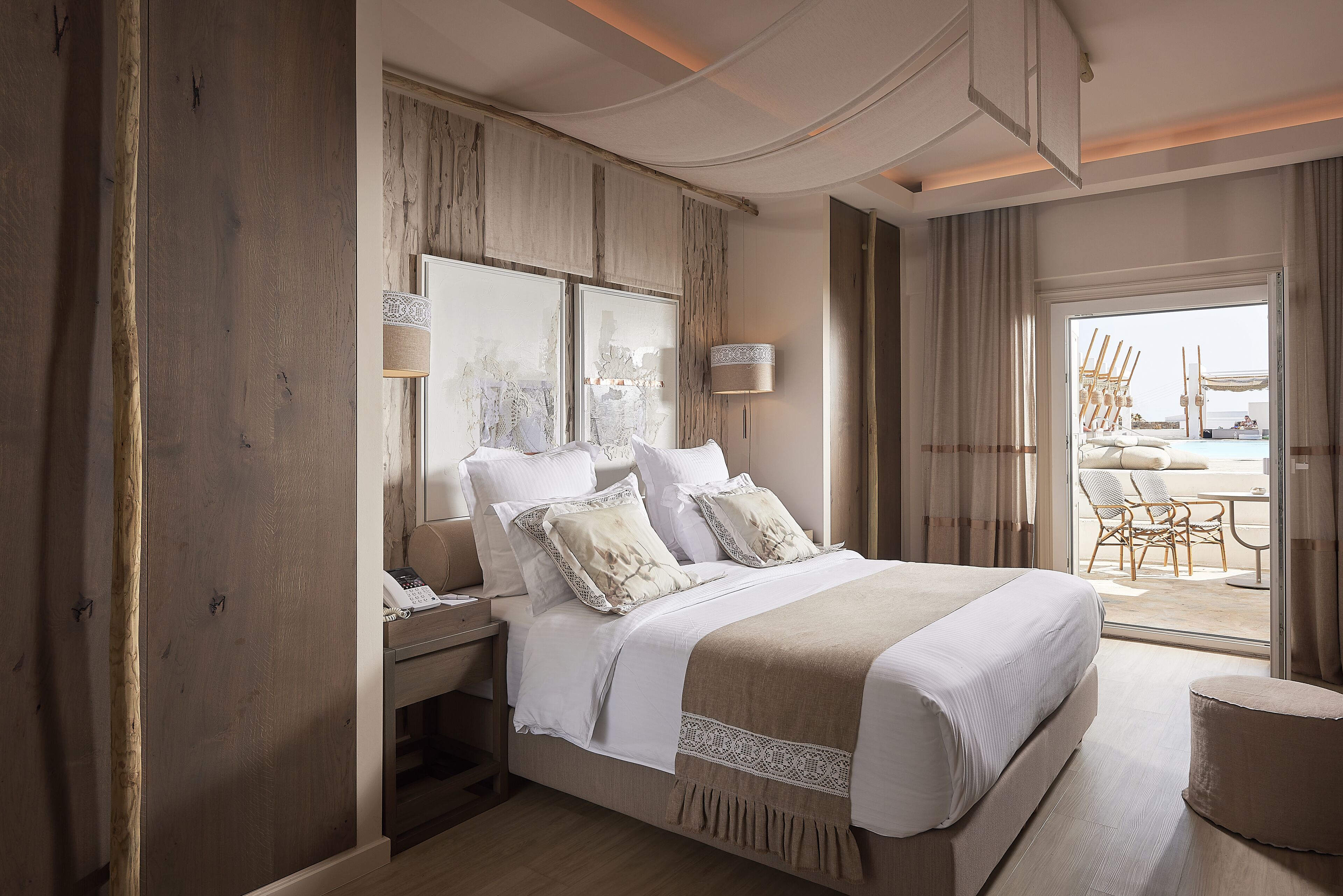 A modern hotel room with a large bed, a nightstand, a lamp, and a view of the beach through the window.