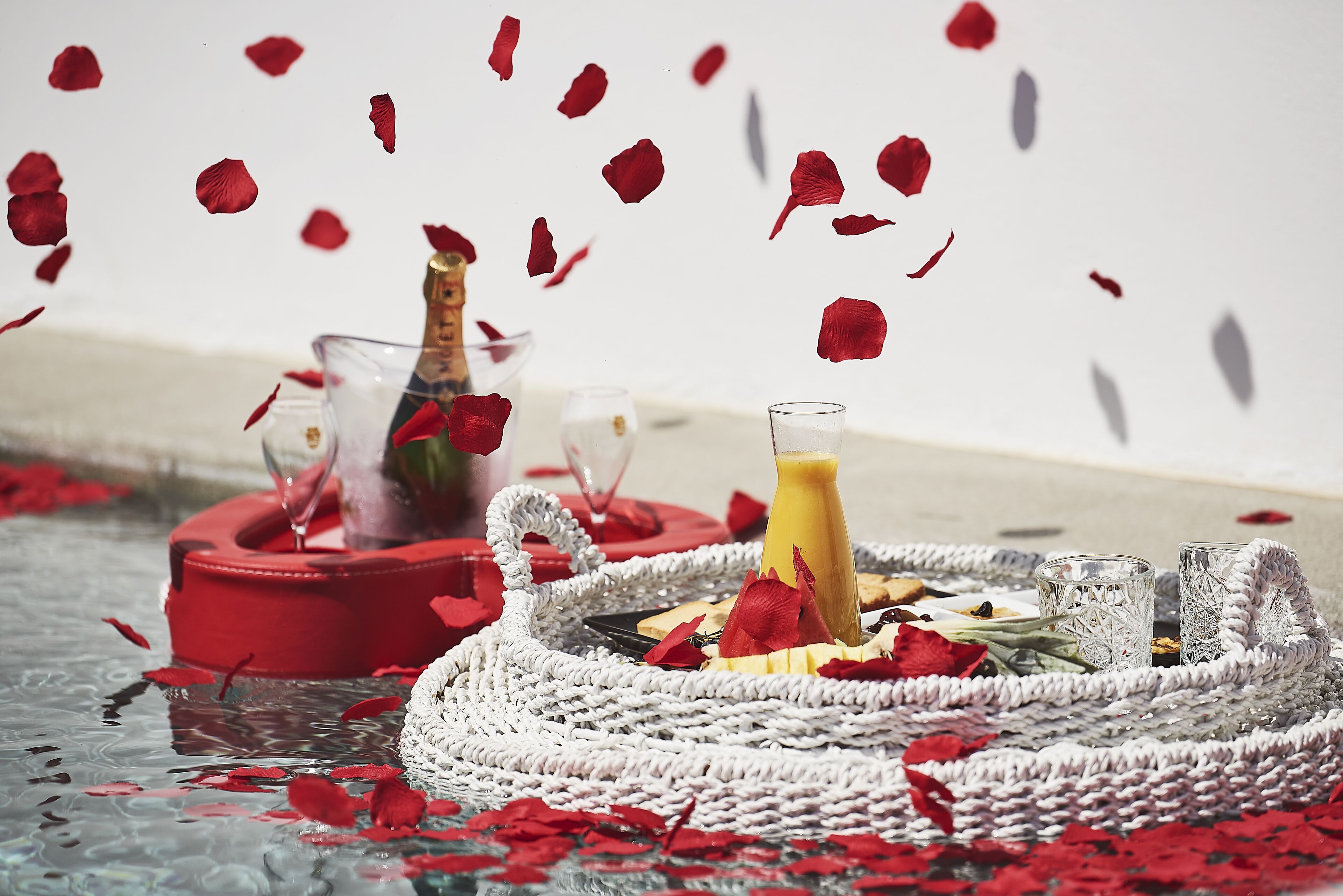 A table with a woven tray, a glass of juice, and a glass of water, surrounded by rose petals.