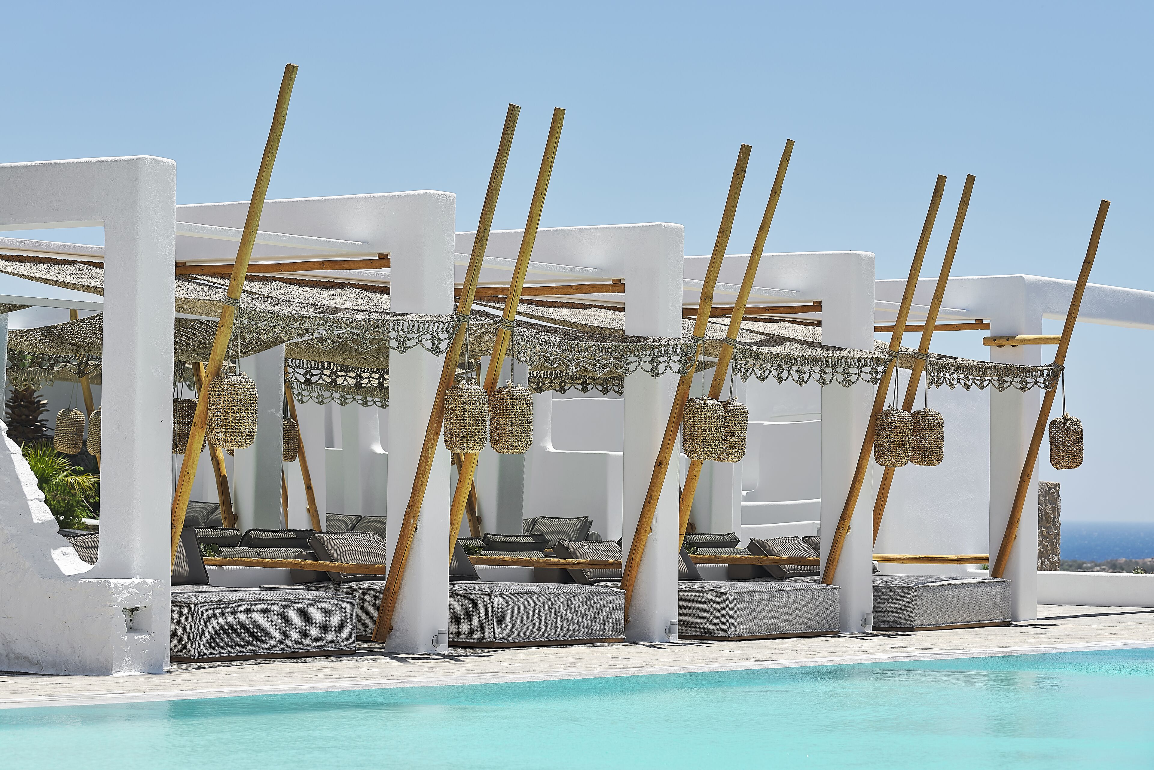 A poolside lounge area with white columns, grey cushioned seating, and woven ceiling decorations.