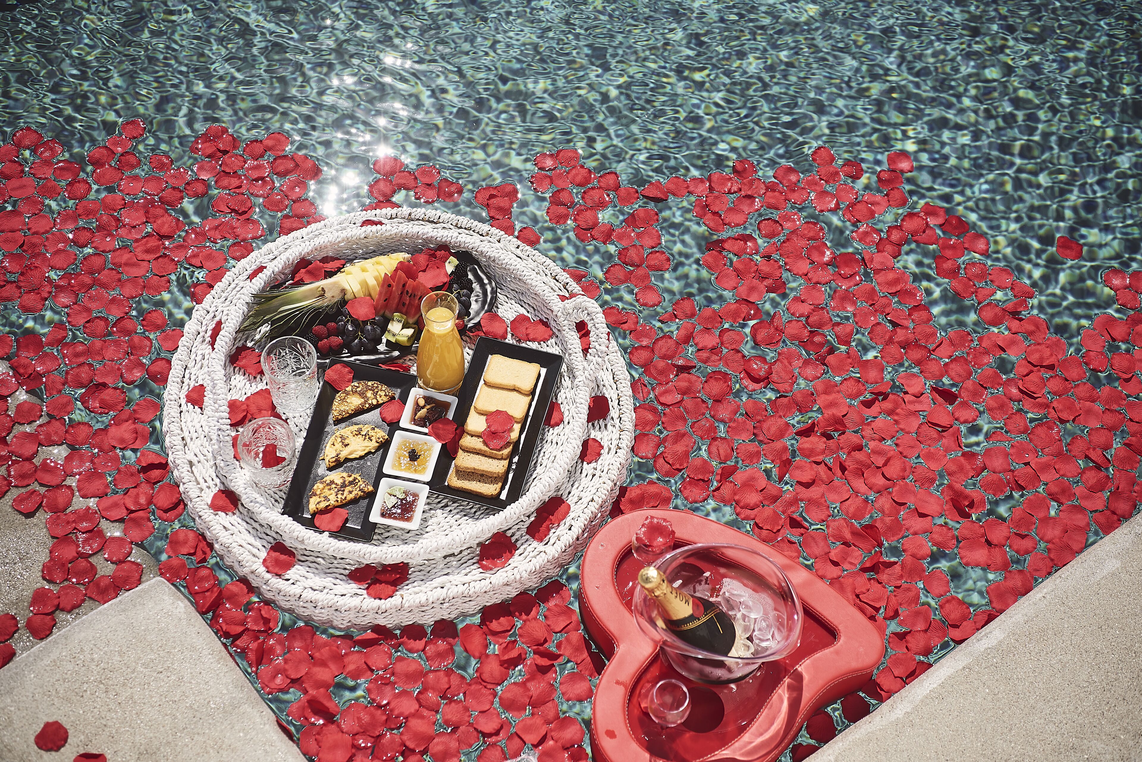 A poolside setup with a basket of food and drinks, surrounded by red rose petals.