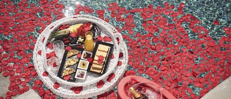 A poolside setup with a basket of food and drinks, surrounded by red rose petals.