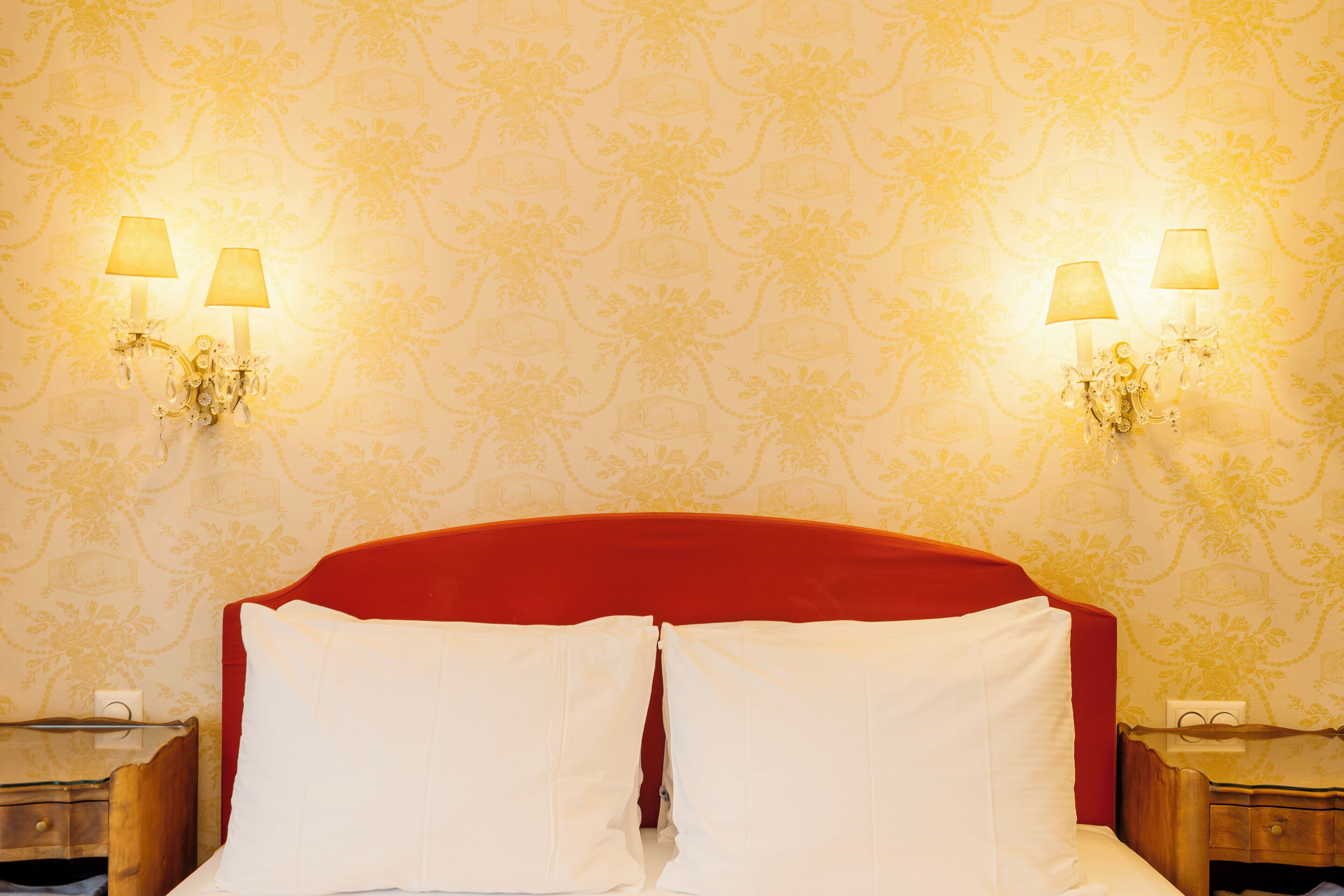 Una habitación de hotel con cabecera roja, dos lámparas de noche y un estampado de flores en el papel tapiz.