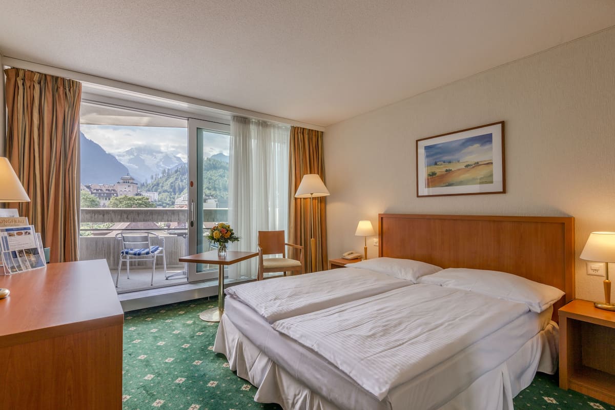Habitación de hotel con dos camas, un escritorio, una silla, una mesa con un florero y vistas a las montañas a través de la ventana.