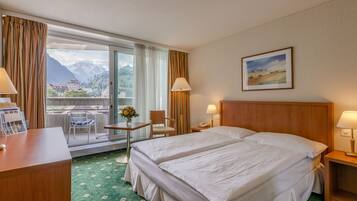 Une chambre d’hôtel avec deux lits, un bureau, une chaise, une table ornée d’un vase de fleurs et une vue sur les montagnes par la fenêtre.