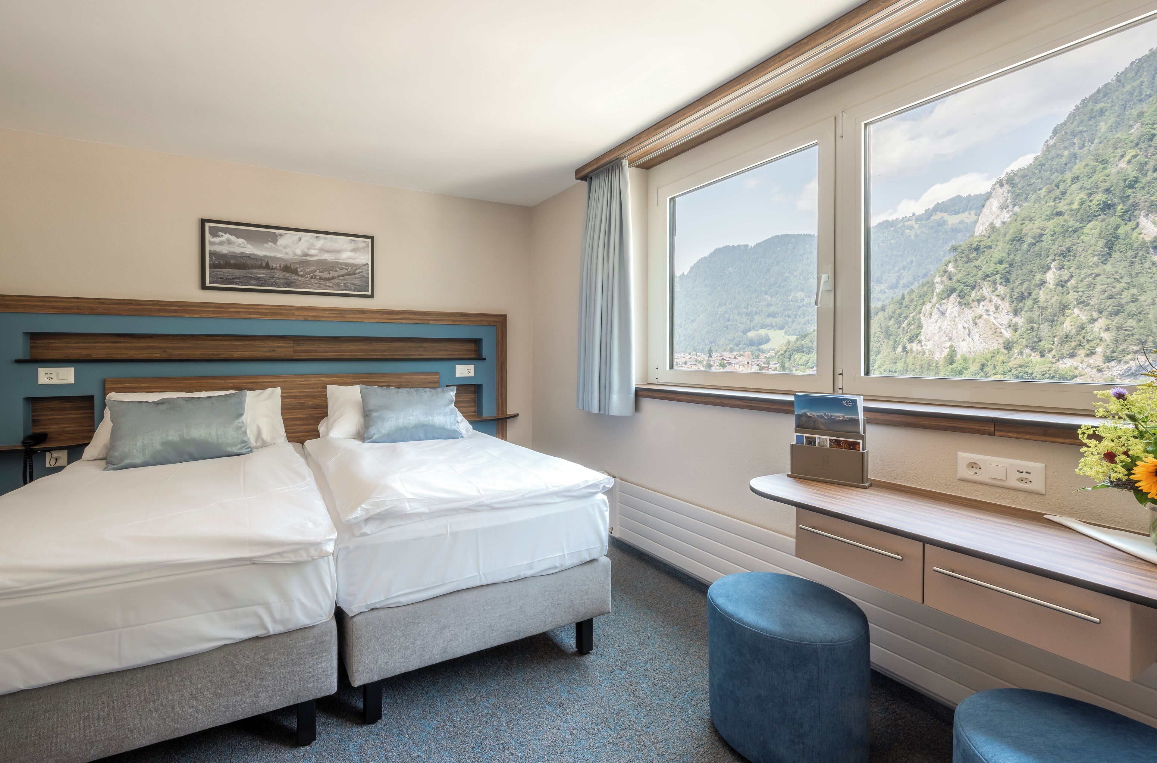 A hotel room with two beds, a desk, a chair, and a window with a mountain view.
