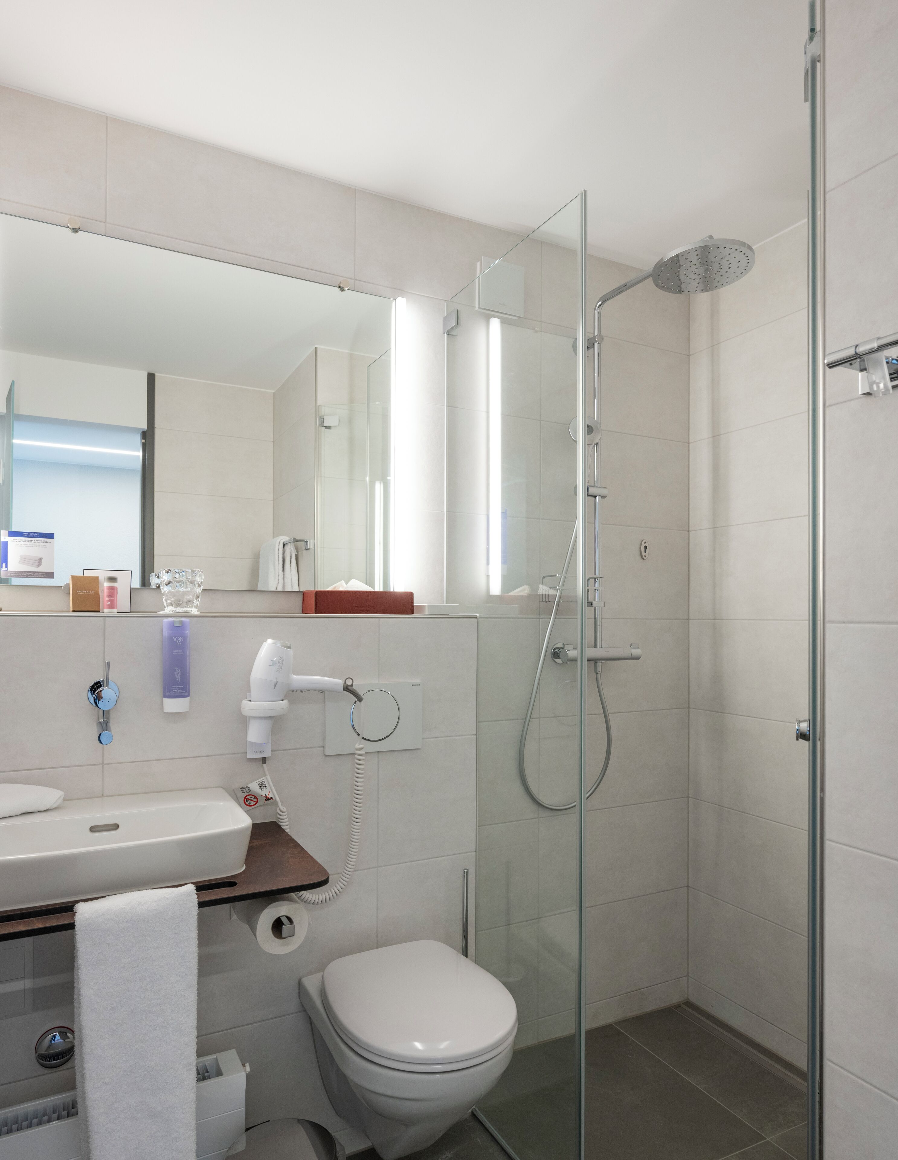Un baño moderno con ducha envuelta en vidrio, inodoro blanco y lavamanos con espejo sobre él.