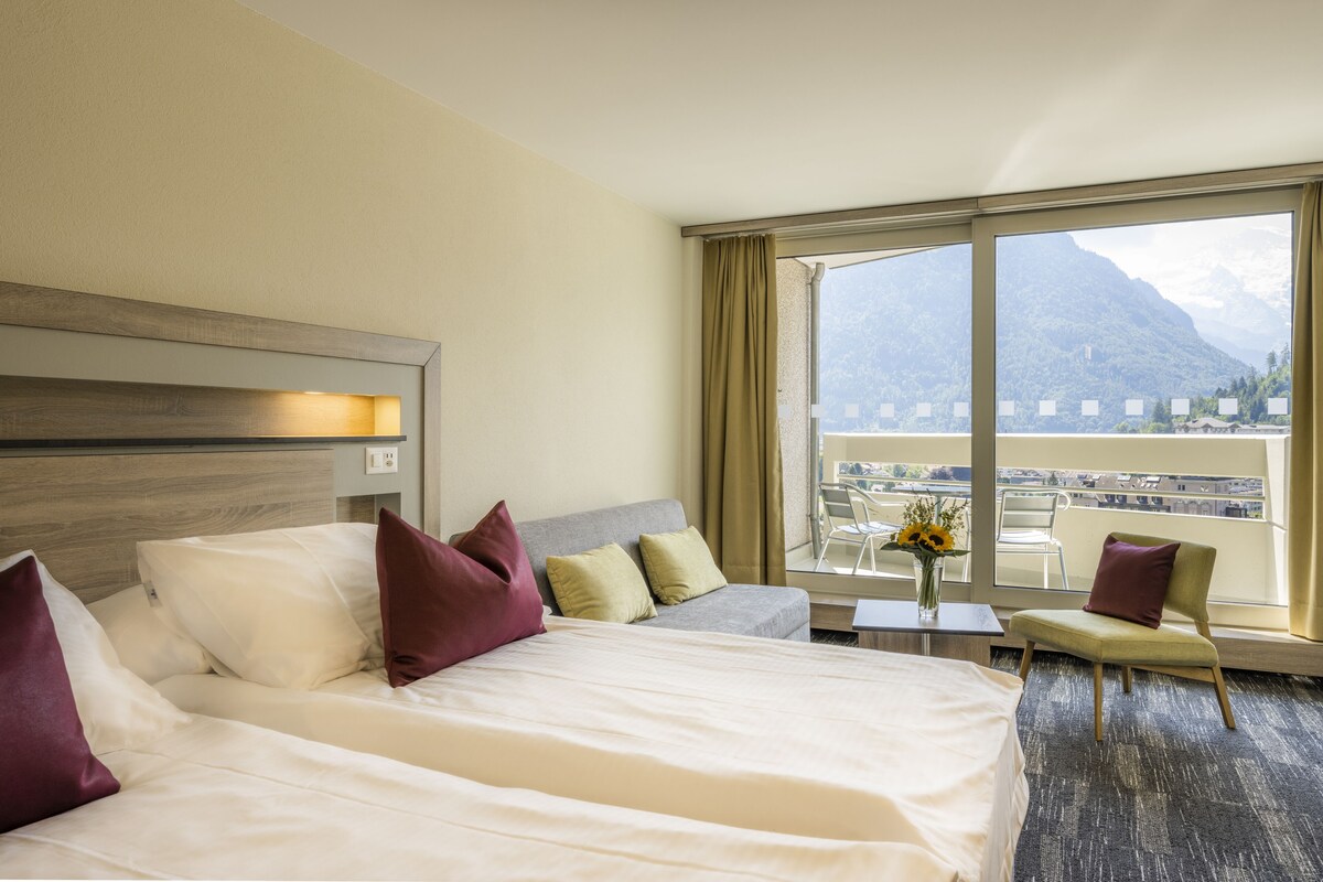 Habitación de hotel con una cama grande, un sofá, una mesita con un jarrón y vistas a montañas a través de amplios ventanales.