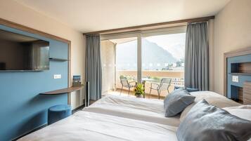 A hotel room with a large bed, a TV mounted on the wall, a balcony with a view of mountains, and a small table with flowers.