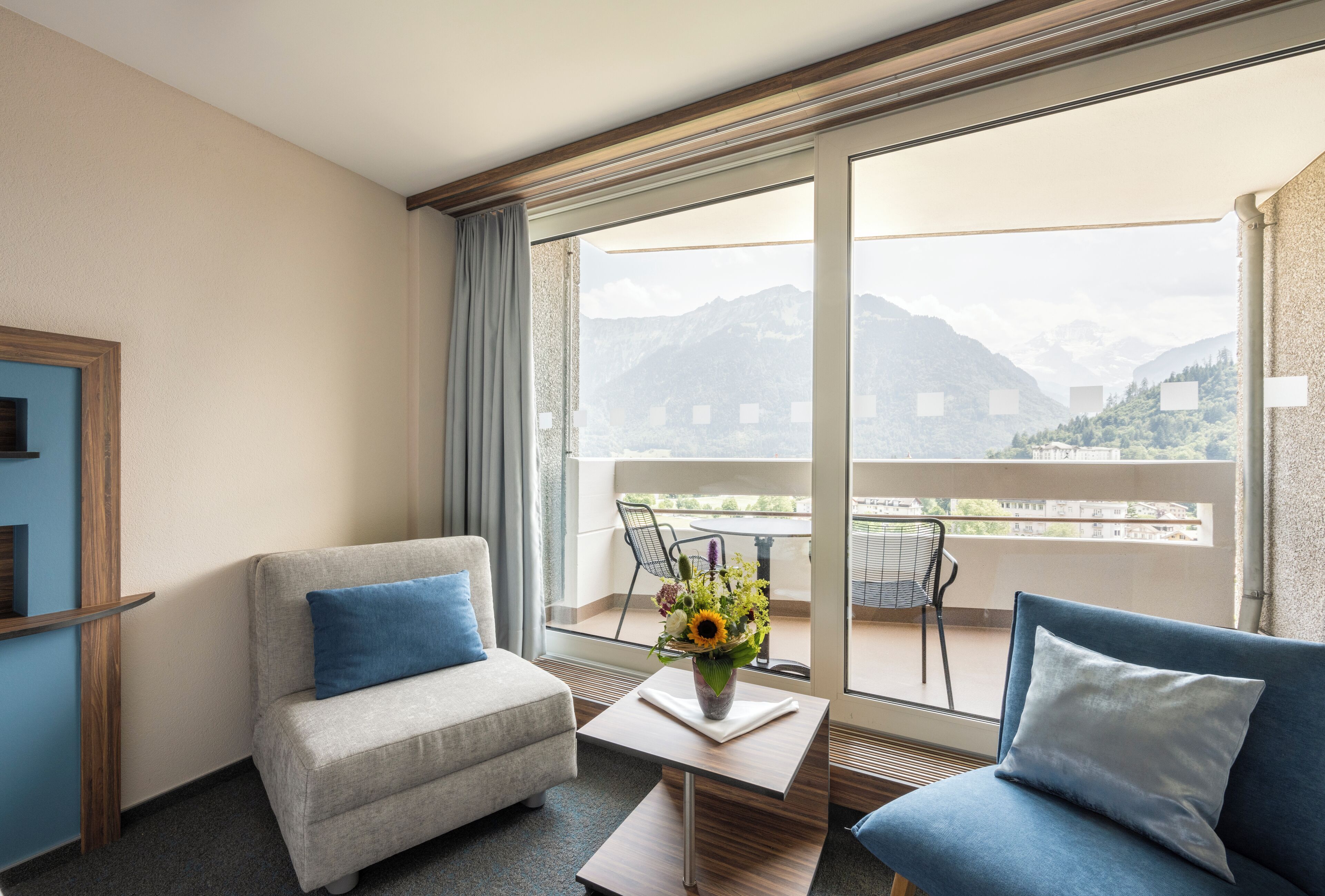 Een hotelkamer met een bank, een stoel, een tafeltje en uitzicht op bergen door grote ramen.