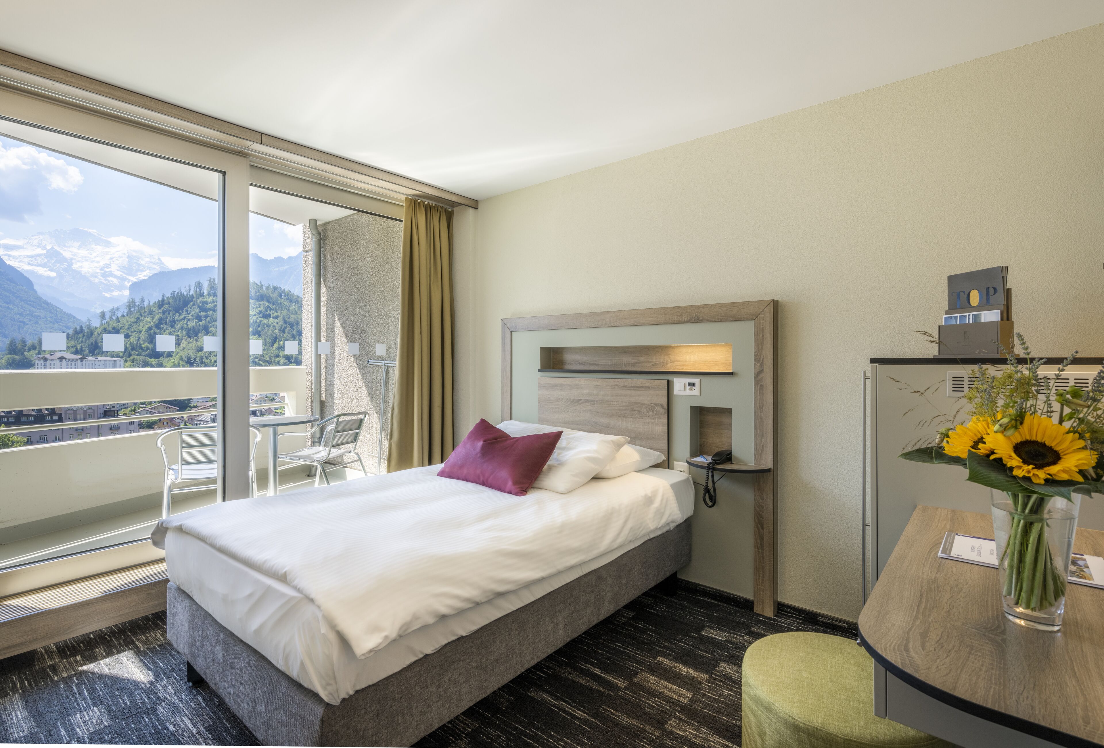 Une chambre d’hôtel avec un lit, un bureau avec un téléphone, une chaise, un vase de fleurs et une vue sur les montagnes, visible à travers de grandes fenêtres.