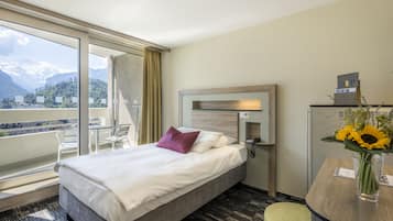 Ein Hotelzimmer mit Bett, Schreibtisch mit Telefon, Stuhl, Blumenvase und Blick auf Berge durch große Fenster.