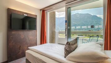 A hotel room with a bed, a flat-screen TV mounted on the wall, and a balcony with a mountain view.