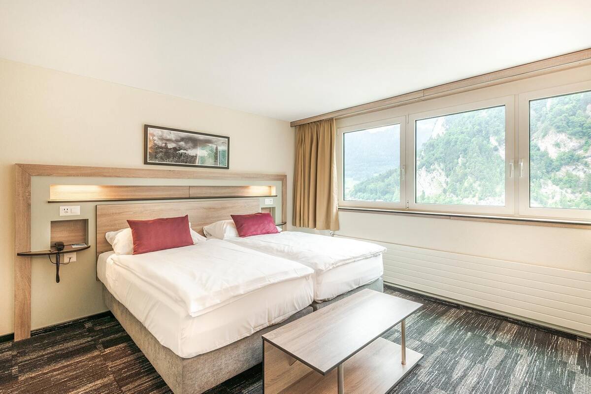 Habitación de hotel con una cama doble, una mesita de noche, un banco, un ventanal con vistas a zonas verdes y un cuadro en la pared.