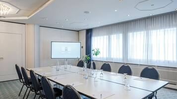 A conference room with a long table, chairs, a projector screen, and a window with curtains.