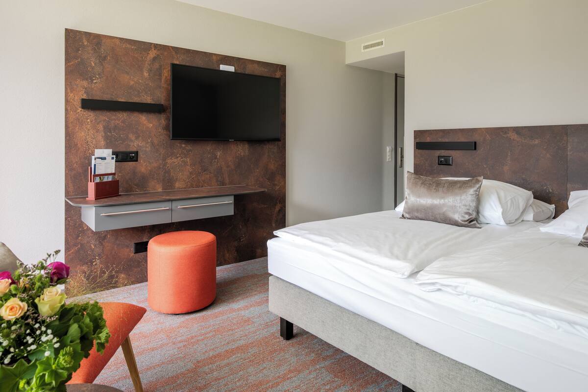 Una habitación de hotel moderna con una cama grande, un televisor montado, un televisor de pantalla plana, una pequeña mesa con un jarrón de flores y un reposapiés rojo.