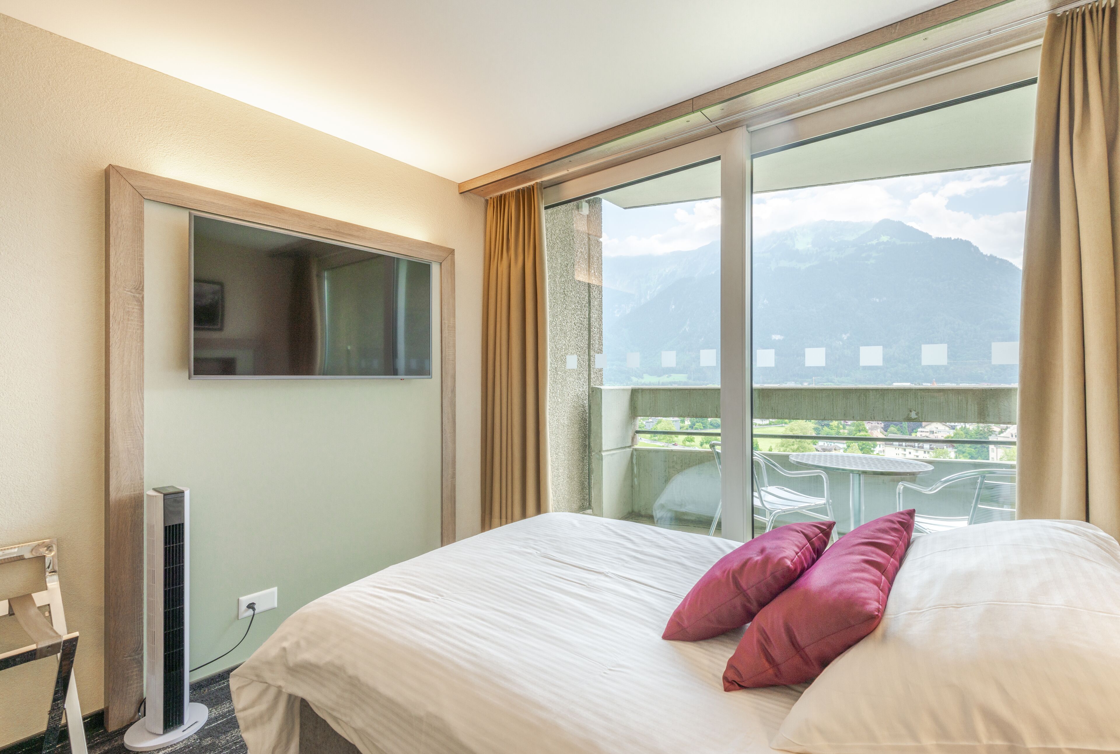 A hotel room with a bed, a TV mounted on the wall, and a view of mountains through large windows.