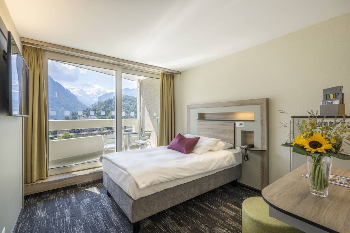Habitación de hotel con cama, escritorio, silla, televisión y un amplio ventanal con vistas a la montaña.