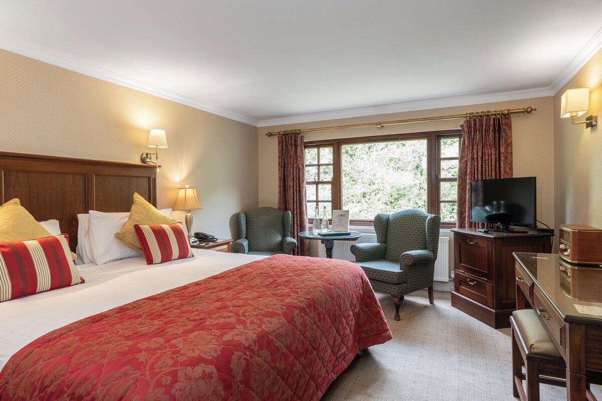 A hotel room with a large bed, two chairs, a desk with a television, and a window with curtains.