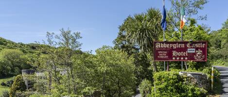 A sign for Abbeville Castle Hotel with a stone wall entrance, flags, and lush greenery.
