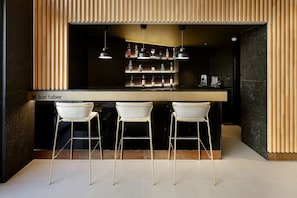A bar area with three white bar stools, a bar counter, and shelves stocked with bottles and glasses. - Hotel HCC Taber (Barcelona)
