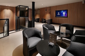A modern lounge area with black leather chairs, a round table with a menu, and a television displaying the Champions League logo. - Hotel HCC Taber (Barcelona)