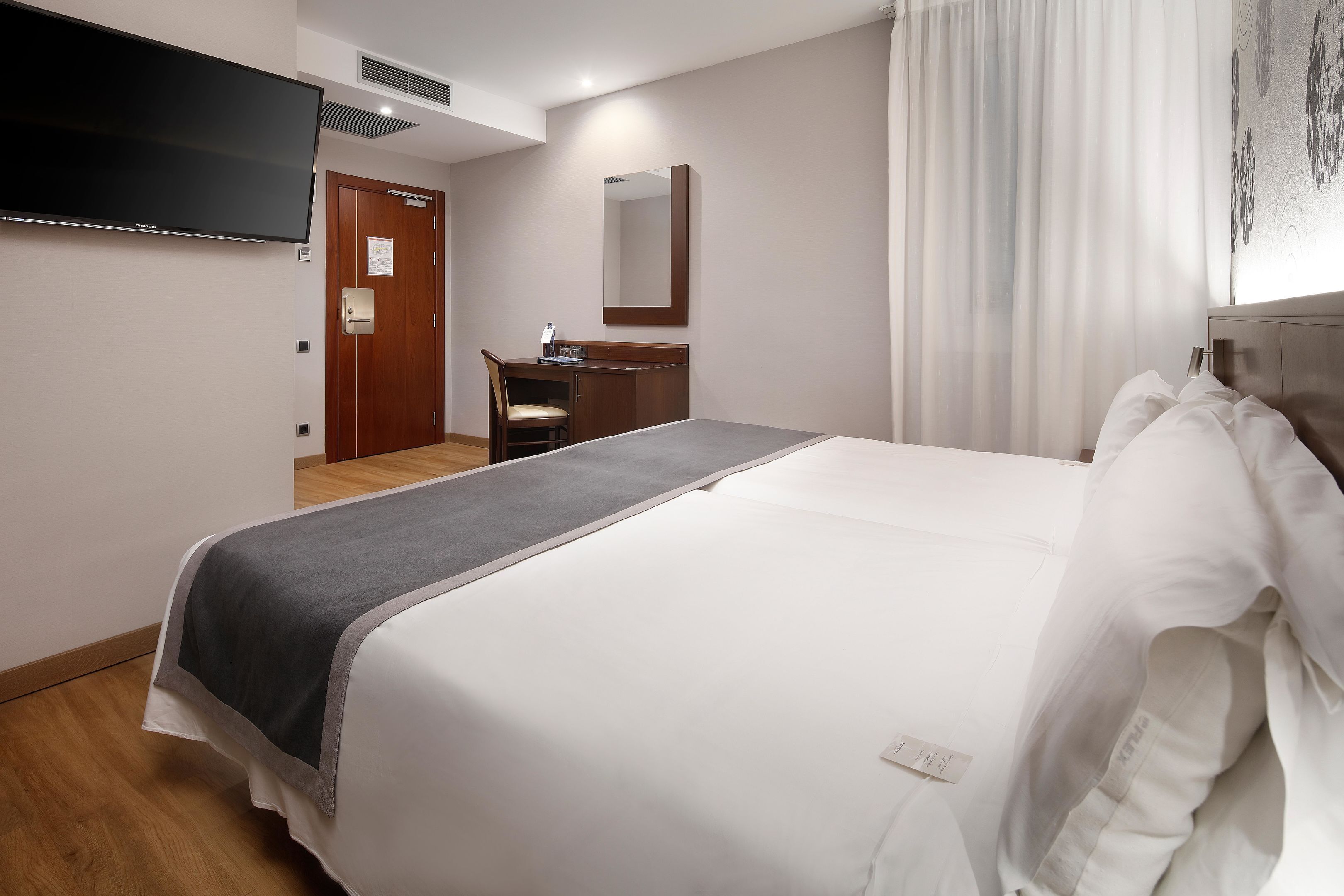 Habitación de hotel con una cama grande, un escritorio con una silla, un televisor de pantalla plana montado en la pared y un espejo sobre el escritorio.