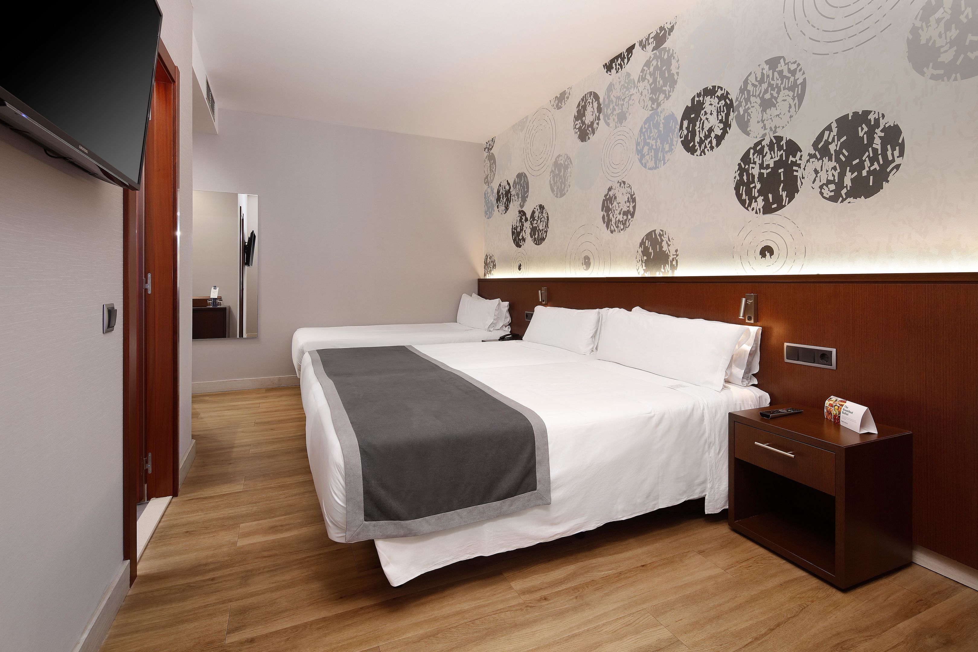Quarto de hotel moderno com cama grande, mesa de cabeceira, TV de tela plana e parede com padrão circular.
