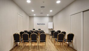 A conference room with rows of chairs, a whiteboard, and a logo for hcchotels Barcelona. - Hotel HCC Taber (Barcelona)