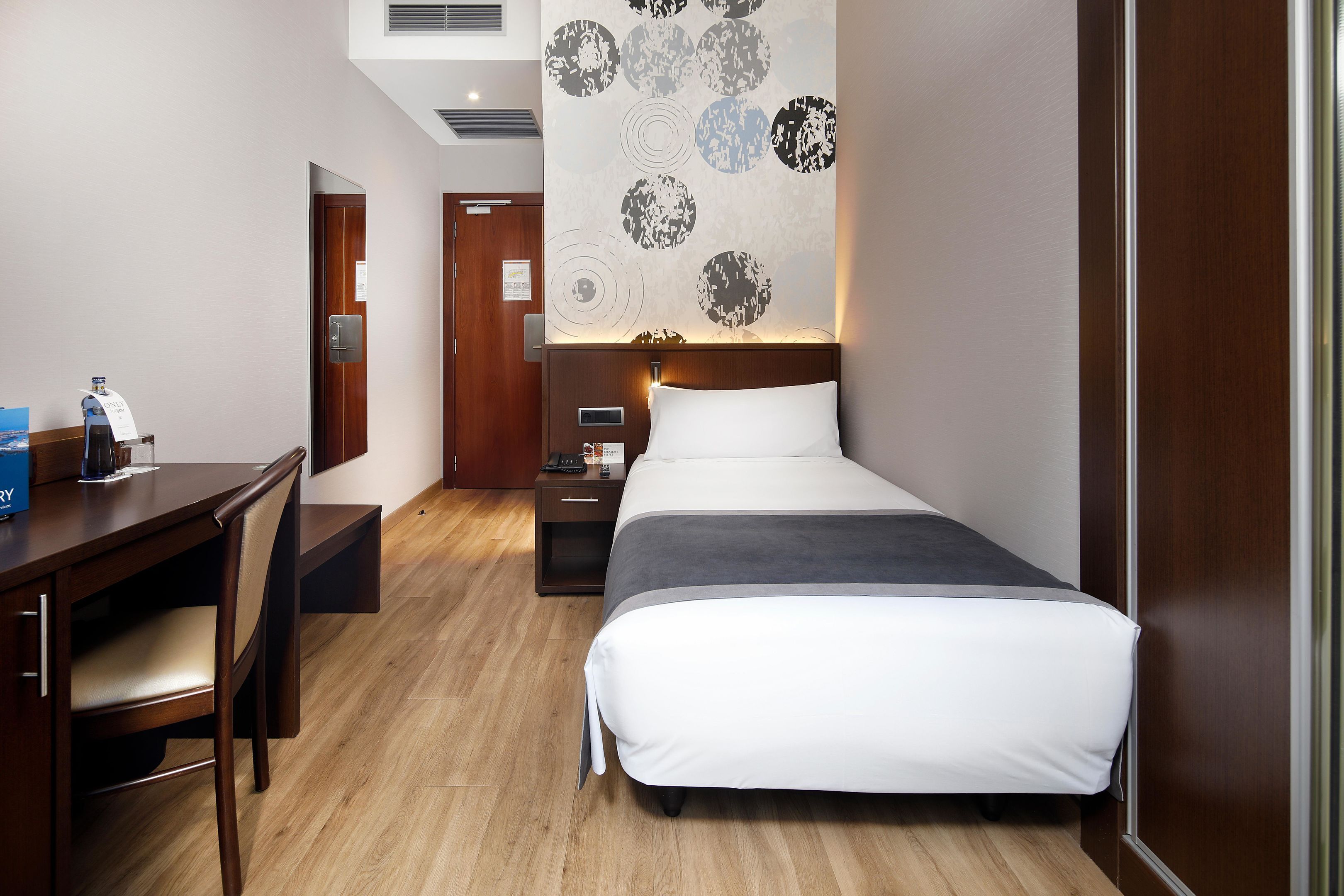 Hotelkamer met een bed, een bureau en een stoel. Er is een wand met cirkelvormige patronen en een nachtkastje met een lamp.