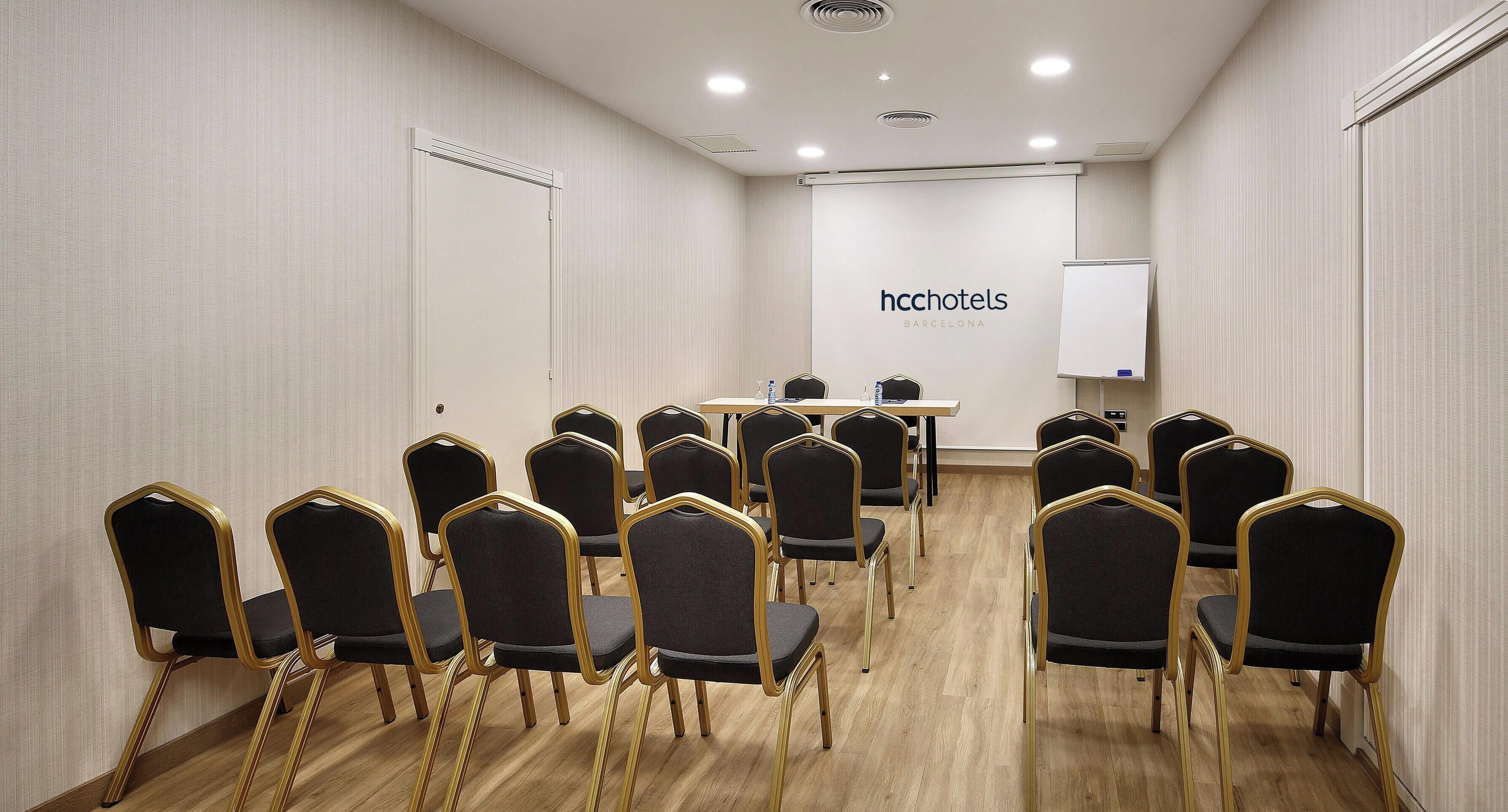 Un salón de conferencias con filas de sillas, una mesa con botellas de agua y una pizarra con el texto 'hchotels'.