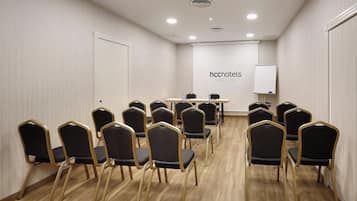 Un salón de conferencias con filas de sillas, una mesa con botellas de agua y una pizarra con el texto 'hchotels'.