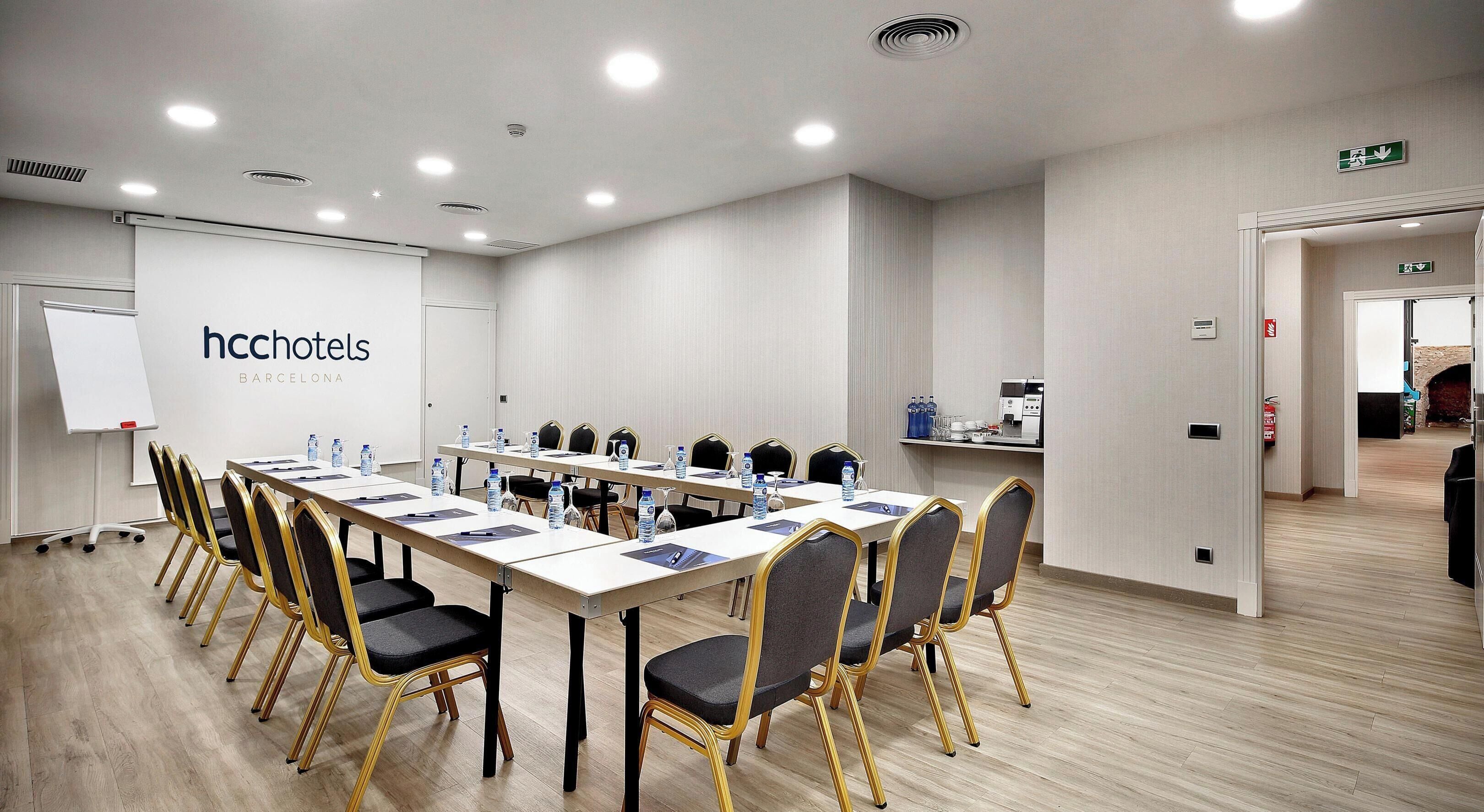 Un salón de conferencias con una larga mesa, sillas, una pizarra y un cartel de HccHotels Barcelona.
