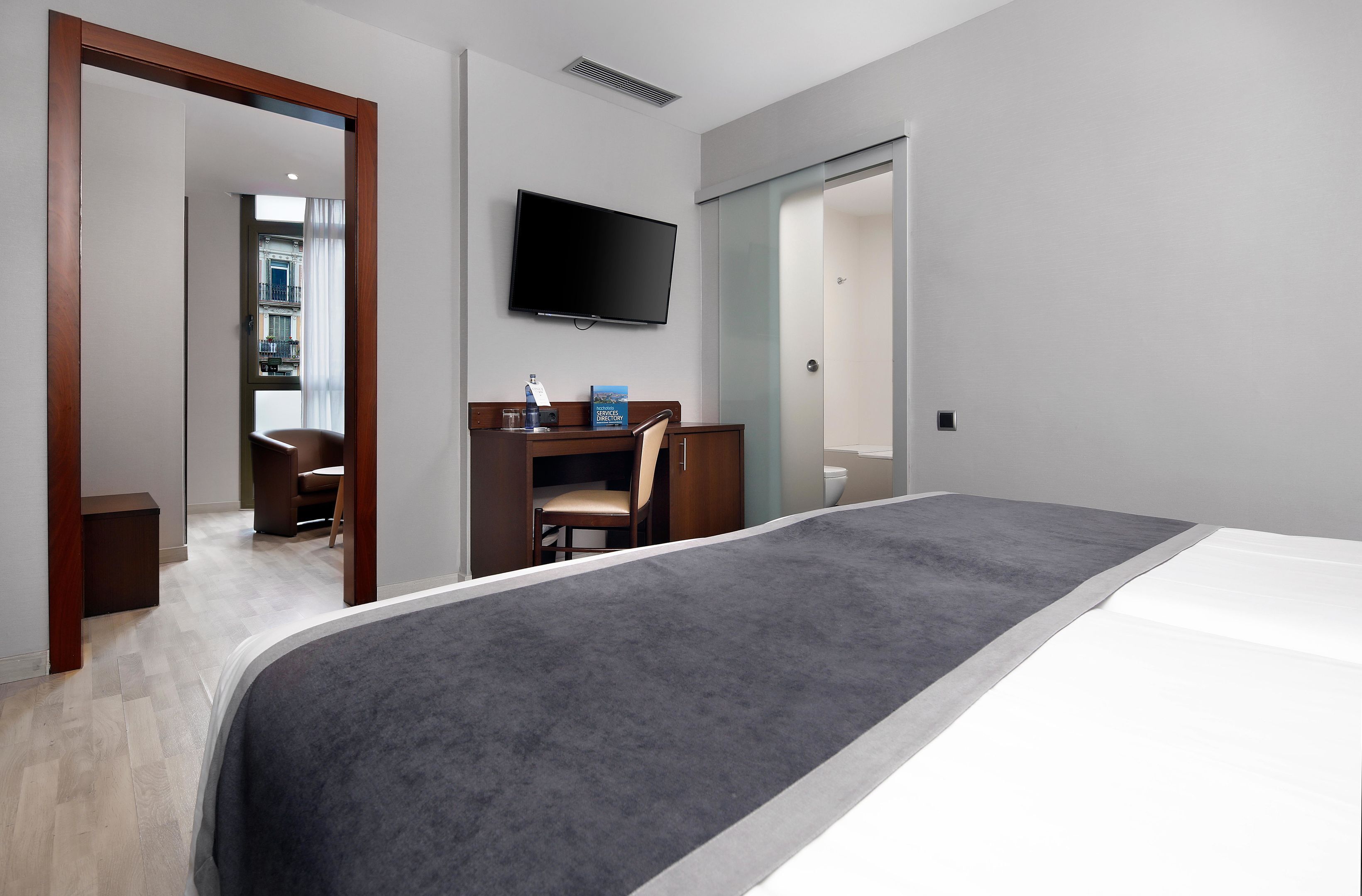 Ein Hotelzimmer mit einem großen Bett, einem Schreibtisch mit Stuhl, einem an der Wand befestigten Fernseher und einem durchgehenden Türspalt einsehbaren Badezimmer.