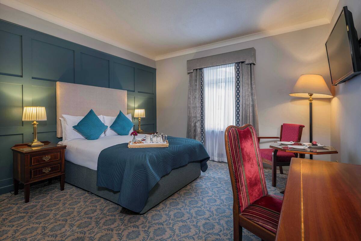 A hotel room with a large bed, bedside tables, a desk, a chair, and a TV mounted on the wall.