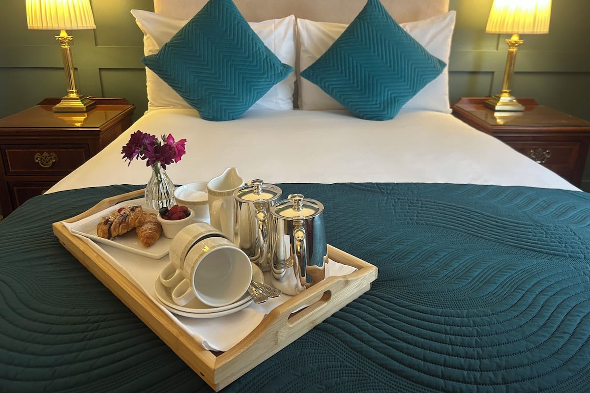 A neatly made bed with teal bedding, two matching pillows, and a tray with breakfast items including croissants, coffee cups, and a vase of flowers.