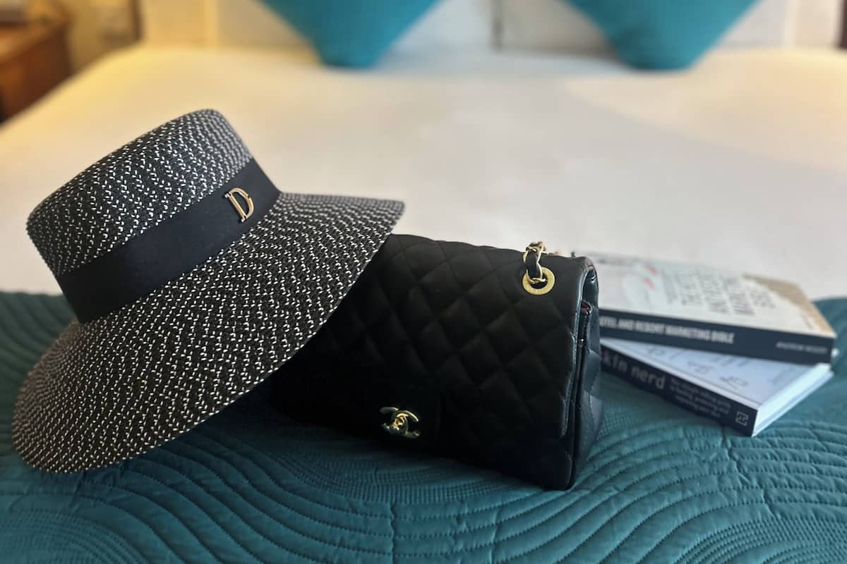 A bed with a teal comforter, two pillows, a black quilted handbag, a black hat, and a pair of glasses on the bed.