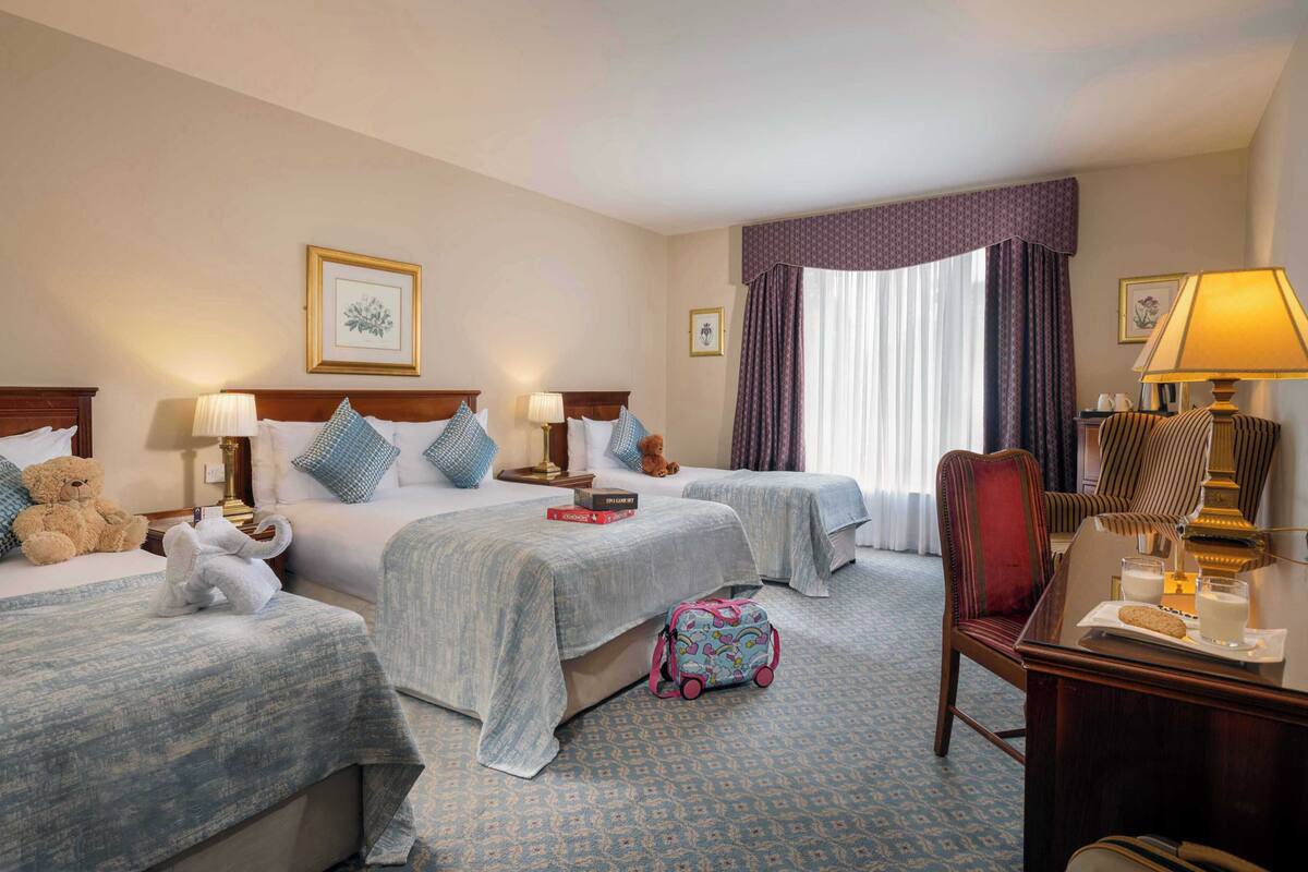 A hotel room with two beds, a desk, a chair, a lamp, and a window with curtains.