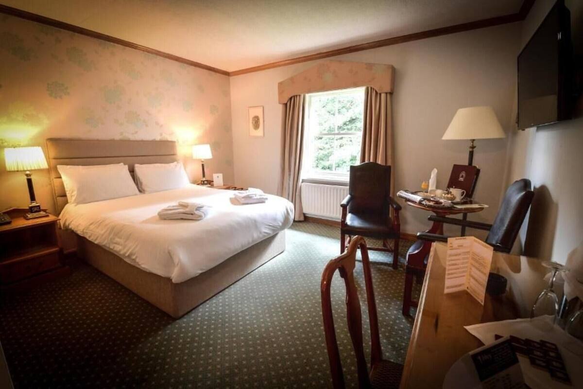A hotel room with a bed, a desk with a chair, a window with curtains, and a television mounted on the wall.