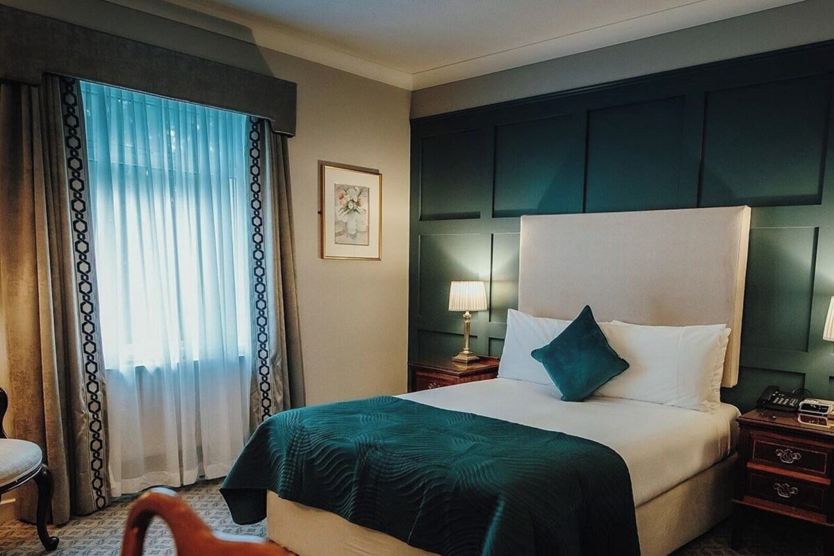 A hotel room with a large bed, two bedside tables with lamps, a chair, and a window with curtains.
