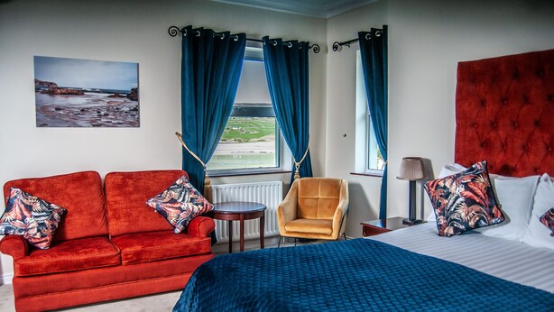 Una habitación con un sofá rojo, un sillón amarillo, una cama con colcha azul y vistas al mar a través de una ventana con cortinas azules.