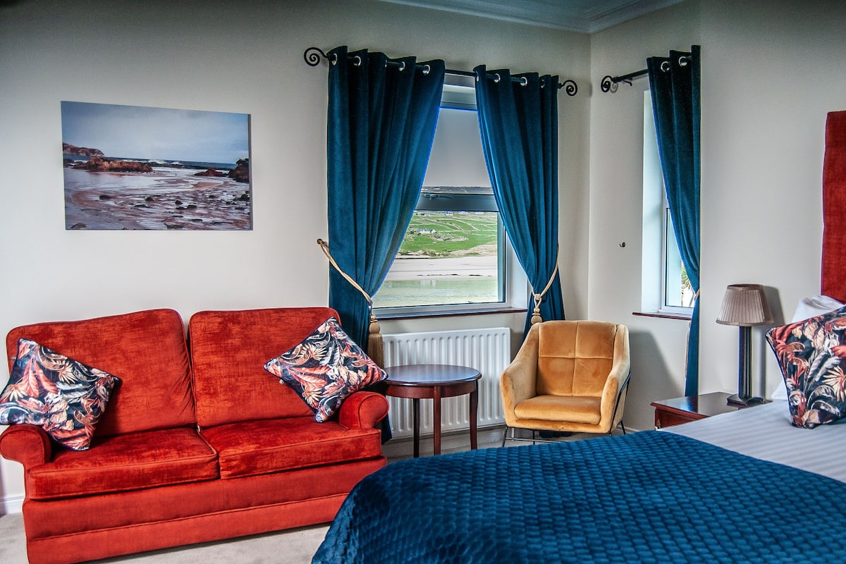 Une pièce avec un canapé rouge, un fauteuil jaune, un lit recouvert d’une couverture bleue et une vue sur la mer par une fenêtre ornée de rideaux bleus.
