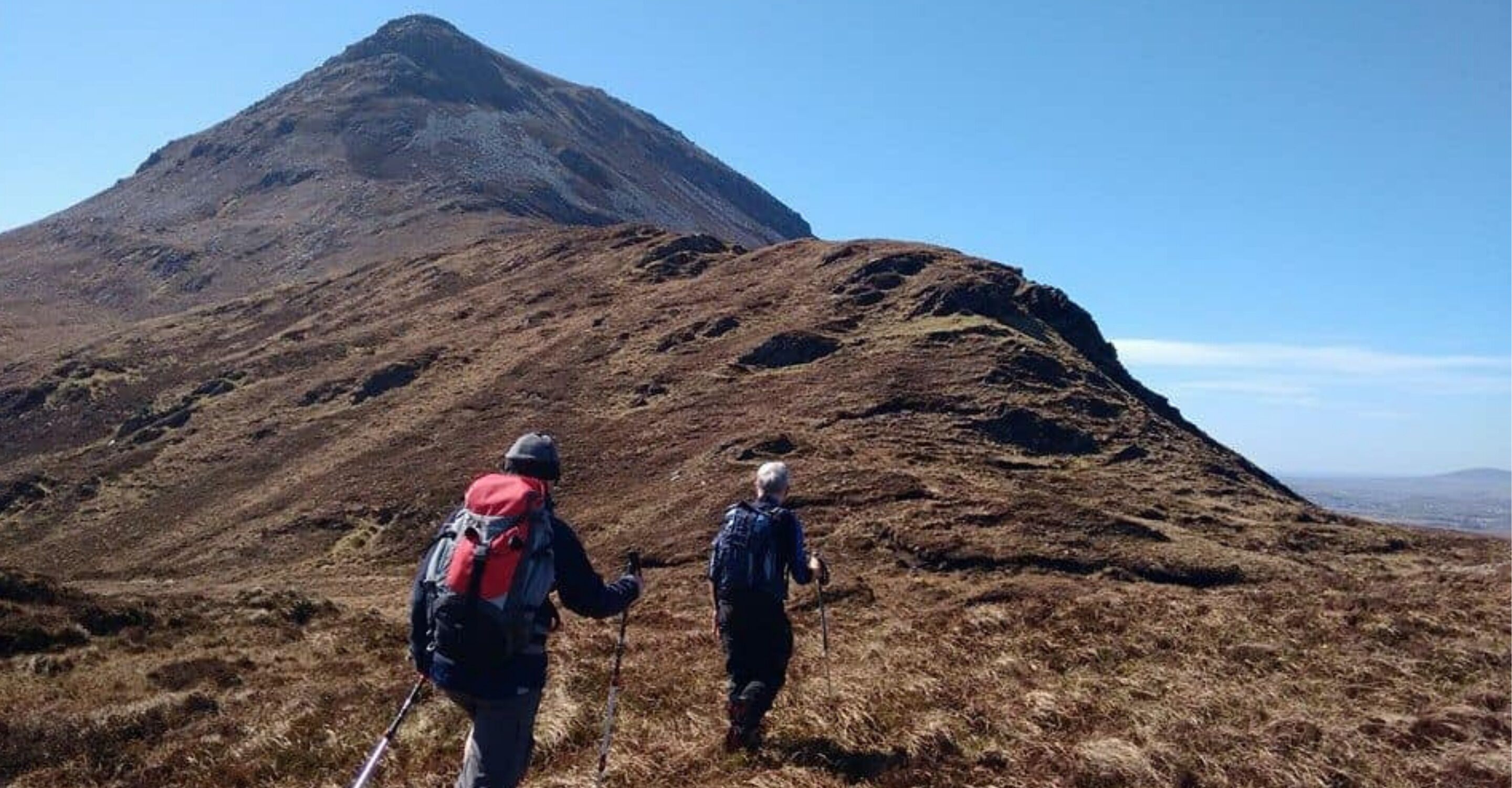 Two hikers on a mountainous trail with a prominent peak in the background.